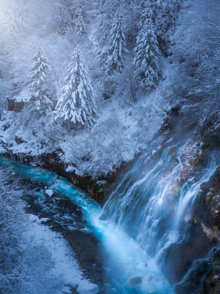 [画像1]美瑛町白髭の滝厳冬期の朝、凍りついた木々と岩の間を流れおちるち青い川へと冬ならではの美しい寒さです