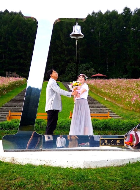 [画像1]太陽の丘えんがる公園コスモス園のオブジェ『Lの鐘』は、LOVEの「L」をモチーフにした『愛の鐘』です。恋人やご夫婦、またご家族でぜひ『Lの鐘』で写真を撮って下さい。この写真はこれからご結婚をされるカッ
