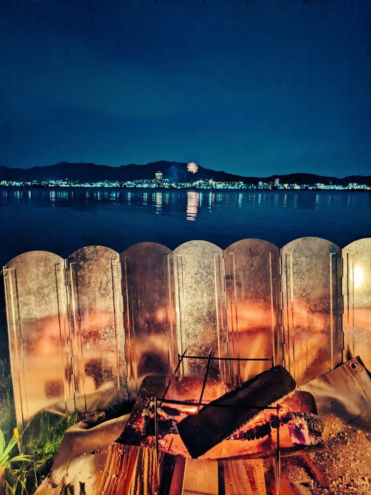 [画像1]琵琶湖畔ソロキャンプでの一枚です。対岸の夜景と焚き火の灯りが幻想的です。花火が水面を照らし、なんとも言えぬ美しさです。